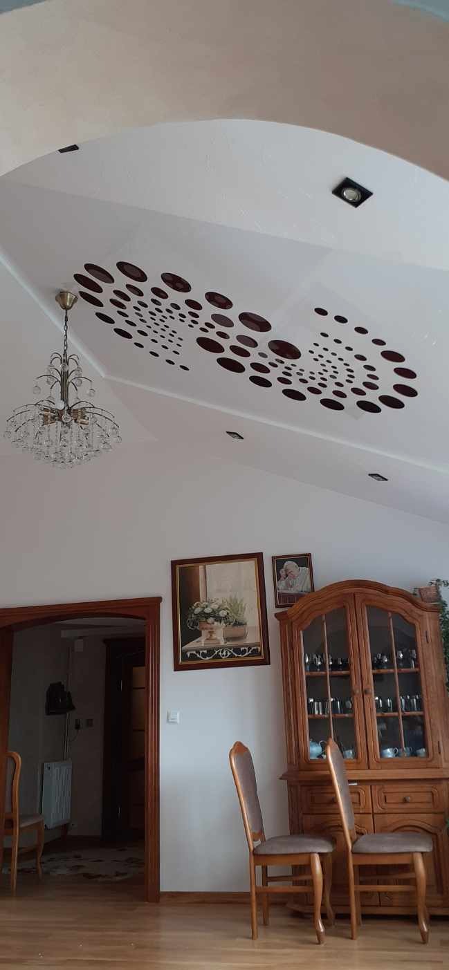 Pokój dzienny z białym sufitem podwieszanym, na którym widnieje wzór z brązowych okręgów o różnej wielkości, kryształowy żyrandol, drewniane meble i obrazy na ścianie.
