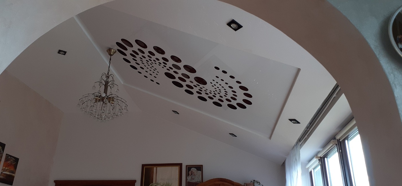 Biały sufit podwieszany z geometrycznym wzorem z brązowych okręgów, oświetlenie punktowe i kryształowy żyrandol w przestronnym wnętrzu z łukowym przejściem.