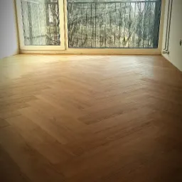 Salon z nową podłogą drewnianą ułożoną w jodełkę, widok z okna na drzewa bez liści i balkon.