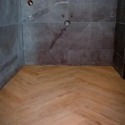 Jasna podłoga ułożona w jodełkę kontrastuje z szarymi, marmurkowymi płytkami ściennymi w łazience z widocznymi otworami na armaturę.