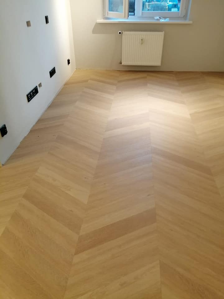 Jasna, dębowa podłoga ułożona we wzór jodełki francuskiej w nowym pomieszczeniu z białymi ścianami i grzejnikiem pod oknem.