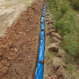 Wykop w ziemi wyłożony niebieską folią, prawdopodobnie pod instalację, widok z góry, obok rosną drzewa iglaste.