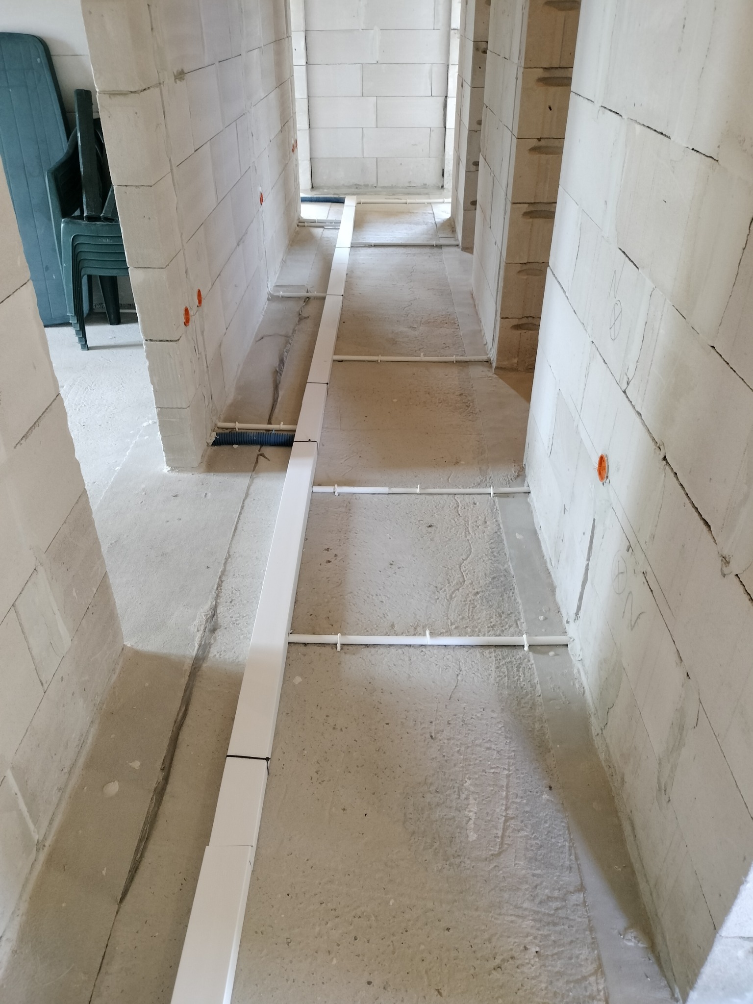 Wnętrze w stanie surowym z widocznymi, białymi korytami kablowymi ułożonymi na podłodze, prowadzącymi wzdłuż ścian z betonu komórkowego. Pomarańczowe puszki elektryczne w ścianach.