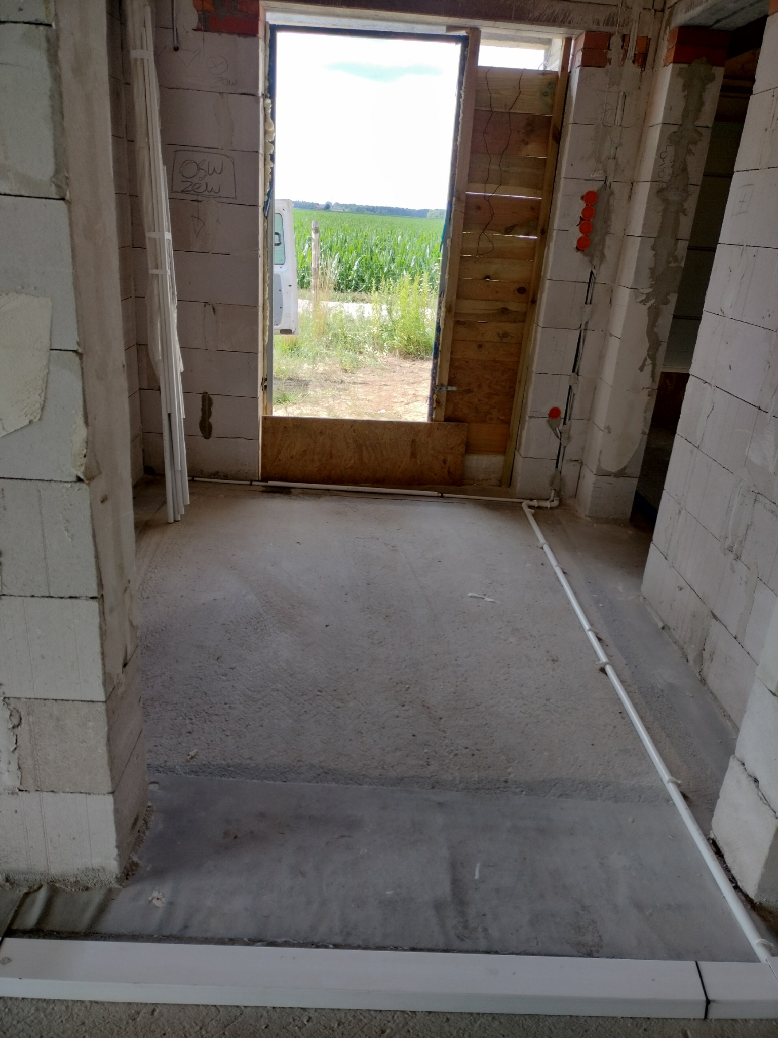 Wnętrze w stanie surowym z widocznymi przewodami elektrycznymi na ścianie, przygotowane do dalszych prac wykończeniowych. Widok na zewnątrz przez otwór drzwiowy.