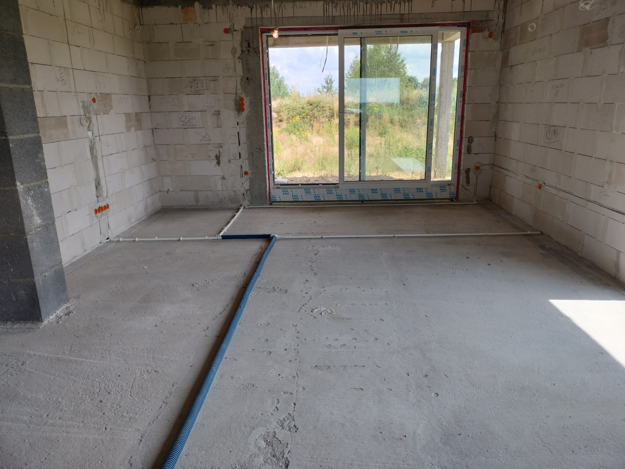 Wnętrze w trakcie instalacji elektrycznej: widoczne kable w peszlach na podłodze surowego pomieszczenia z oknem, ściany z betonu komórkowego z puszkami instalacyjnymi.