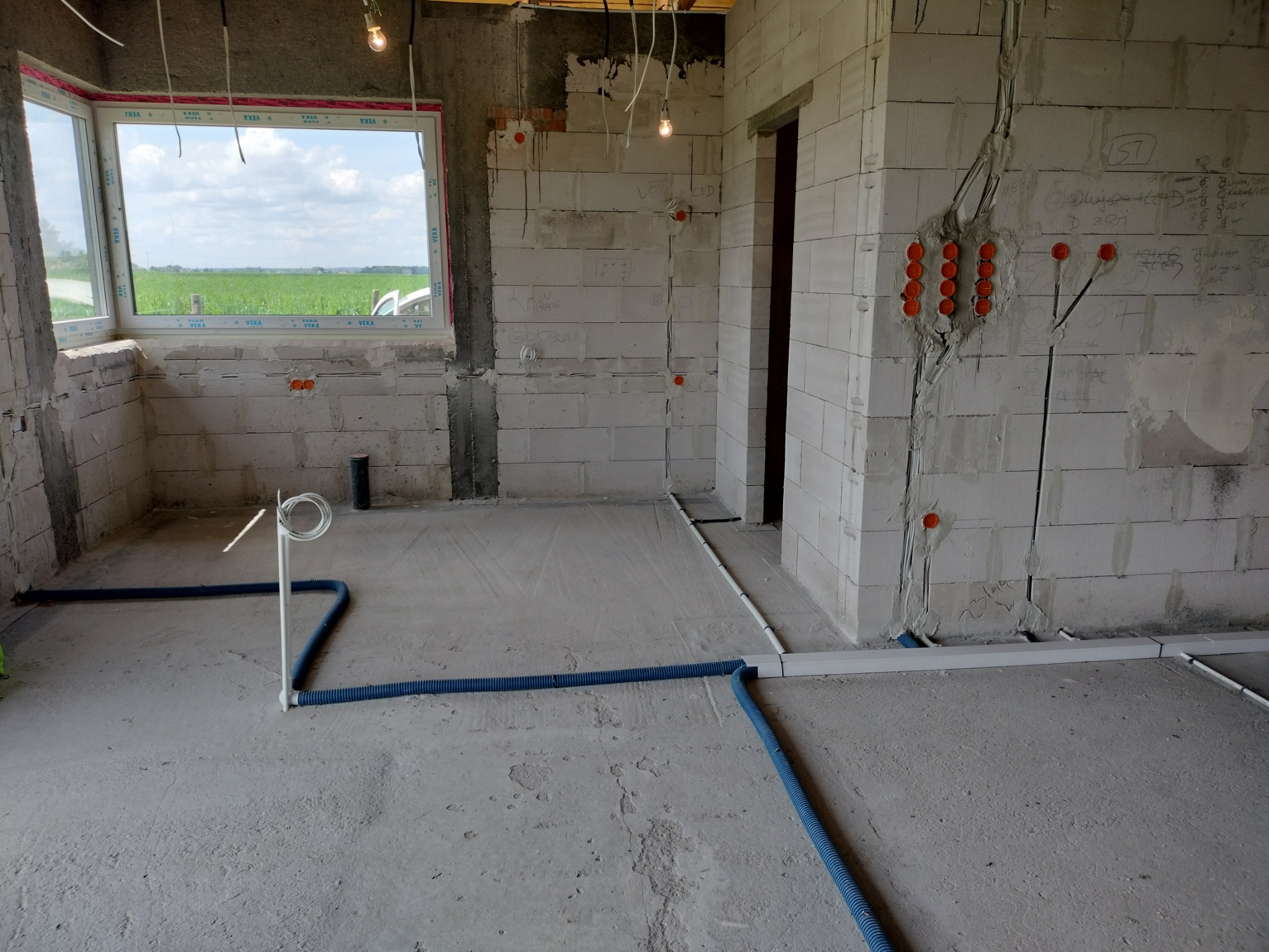 Surowy stan wnętrza z instalacją elektryczną: kable w peszlach, puszki podtynkowe, okno z widokiem na pole. Wnętrze w trakcie prac wykończeniowych.