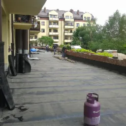 Prace na płaskim dachu budynku wielorodzinnego: robotnicy rozkładają materiał izolacyjny, widoczna butla z gazem, rolki papy, ceglana attyka i otaczająca zieleń.