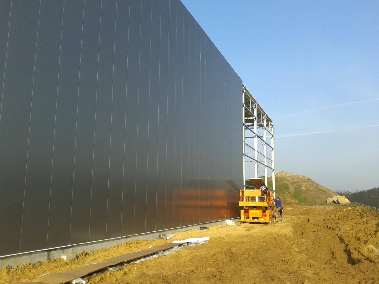 Montaż pionowych płyt warstwowych na stalowej konstrukcji hali, widoczny podnośnik koszowy i teren budowy.