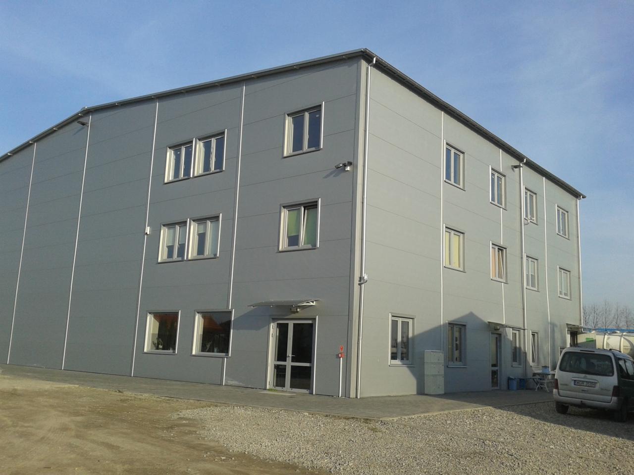 Dwukondygnacyjna hala przemysłowa z szarych płyt warstwowych, widok z zewnątrz w słoneczny dzień, z widocznymi oknami i wejściem.