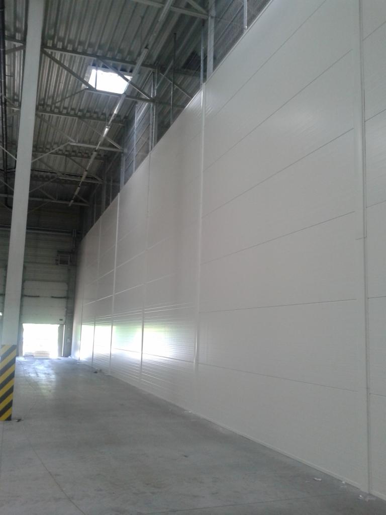 Wnętrze hali produkcyjnej z białymi ścianami z płyt warstwowych, widoczna konstrukcja stalowa dachu i brama wjazdowa na końcu korytarza.