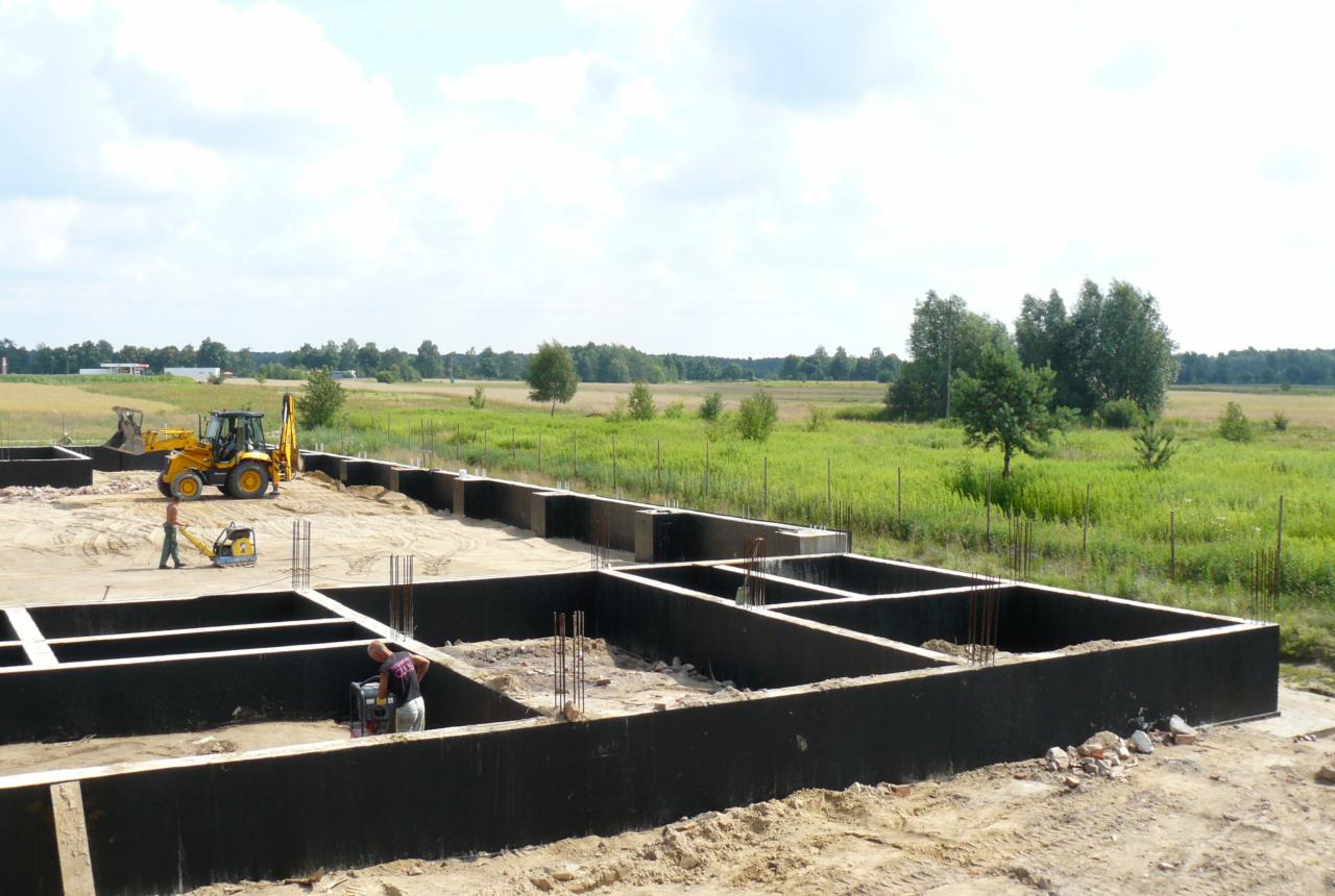 Budowa fundamentów pod halę: widok na wylane fundamenty z wystającymi prętami zbrojeniowymi, na placu budowy pracuje koparko-ładowarka i zagęszczarka, w tle łąka i las.