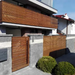 Nowoczesna posesja z drewnianym ogrodzeniem lamelowym i balkonem z identycznym wykończeniem, uzupełniona szarą kostką brukową i dwoma kulistymi krzewami.