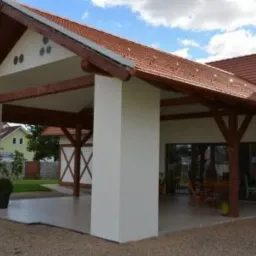 Zadaszony taras z drewnianą konstrukcją nośną i białymi filarami, widok z zewnątrz na meble ogrodowe przez przeszklone ściany.