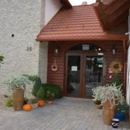 Wejście do budynku z brązowymi drzwiami i zadaszeniem, udekorowane roślinami w donicach i dyniami, z numerem 21 na elewacji.