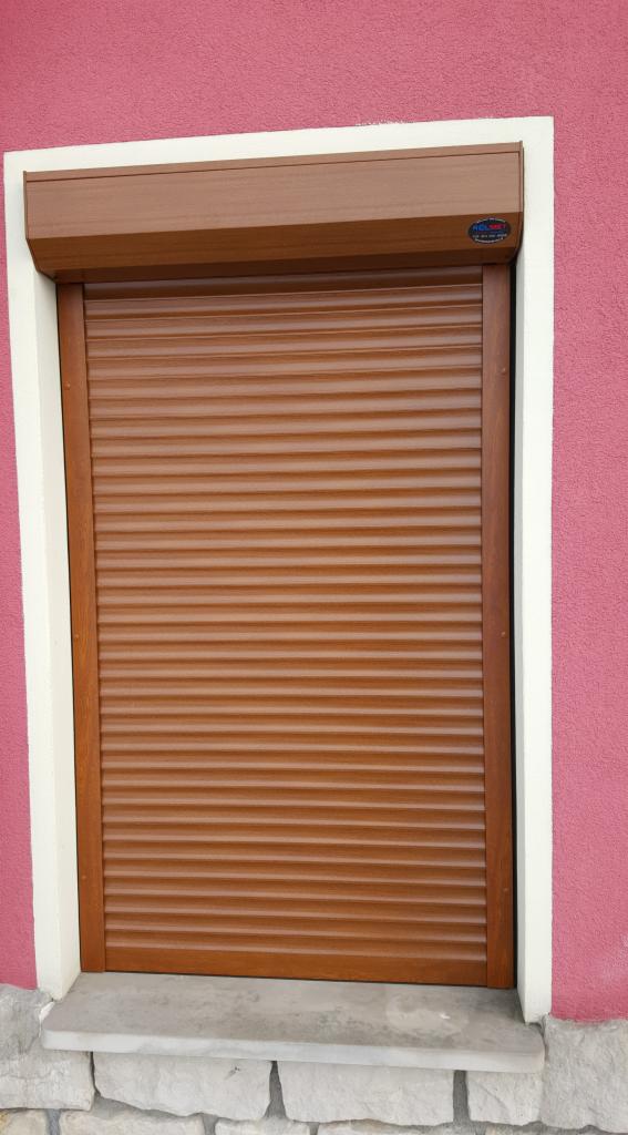 Zewnętrzna roleta okienna w kolorze brązowym, z listwami poziomymi, zamontowana w białej ramie na tle różowej ściany z fakturą.