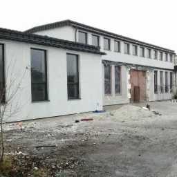 Elewacja budynku hali produkcyjnej 1.2T w trakcie prac wykończeniowych, okna zabezpieczone folią, widoczny stos piasku.