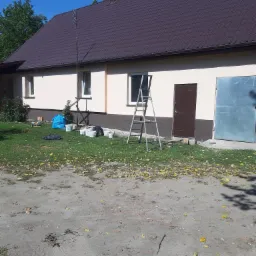 Dom jednorodzinny z beżową elewacją w trakcie prac wykończeniowych; widoczne wiaderka z farbą, drabina i fragment pomalowanej ściany przy drzwiach.