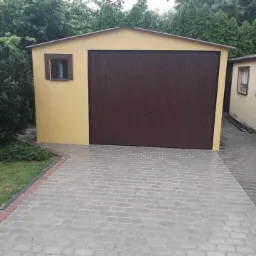 Żółty garaż blaszany z brązową bramą segmentową, widoczny z perspektywy ułożonej kostki brukowej.
