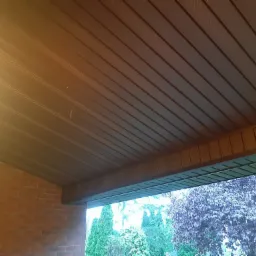 Zadaszenie tarasu z brązowych paneli z widokiem na ogród z ceglaną ścianą po lewej stronie.