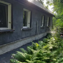 Dom w trakcie prac elewacyjnych, ściany pokryte szarym materiałem izolacyjnym, widoczne białe ramy okienne, otoczenie z bujną roślinnością.