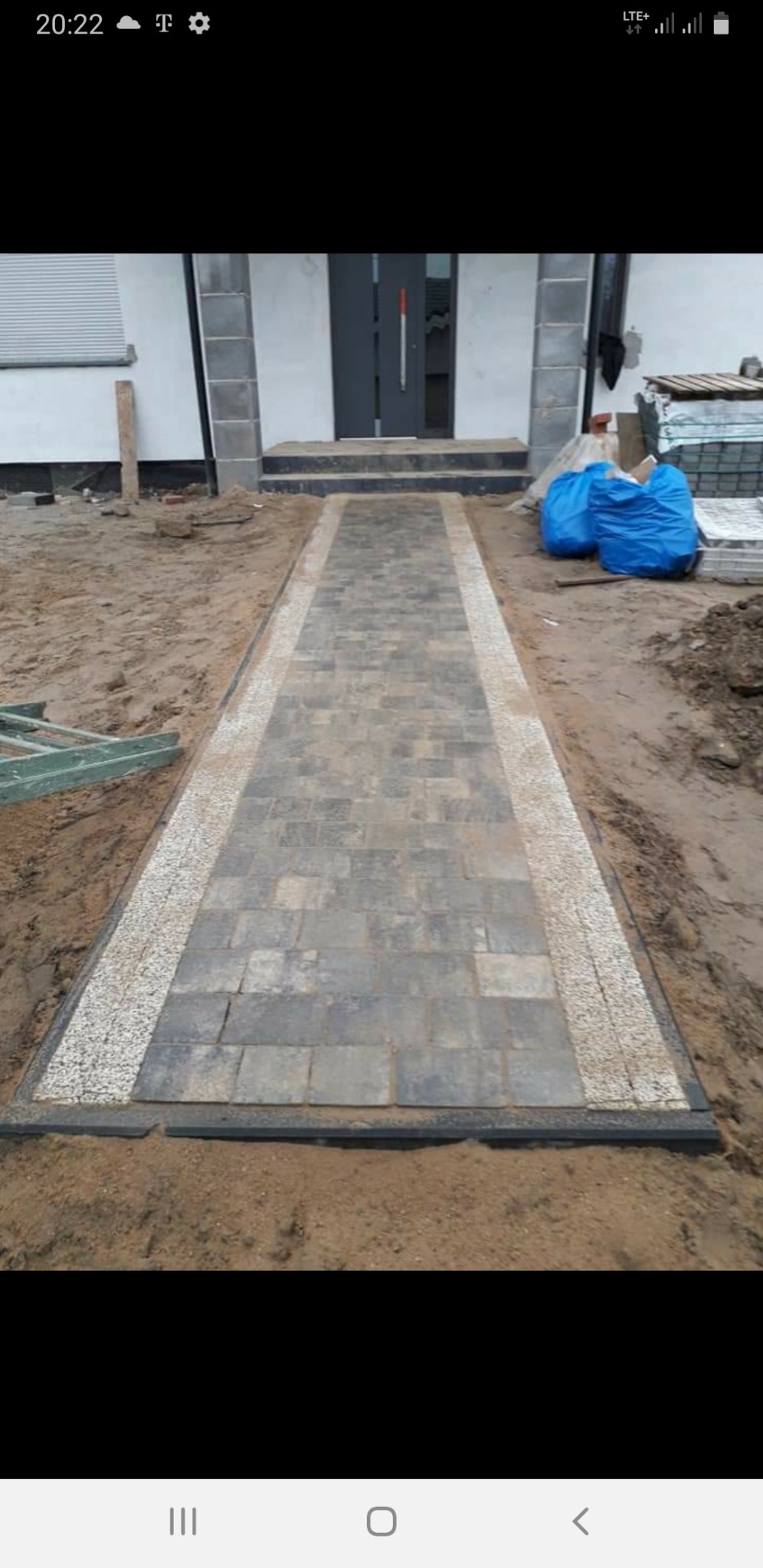Ułożony chodnik z szarej kostki brukowej z obramowaniem z jasnego grysu prowadzący do drzwi wejściowych domu, widoczne worki z materiałami budowlanymi i narzędzia.