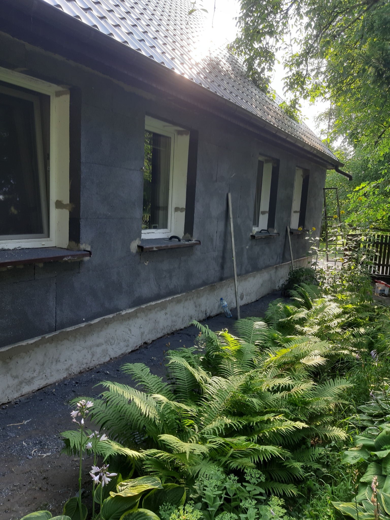 Dom w trakcie prac elewacyjnych, ściany pokryte szarym materiałem izolacyjnym, widoczne białe ramy okienne, otoczenie z bujną roślinnością.