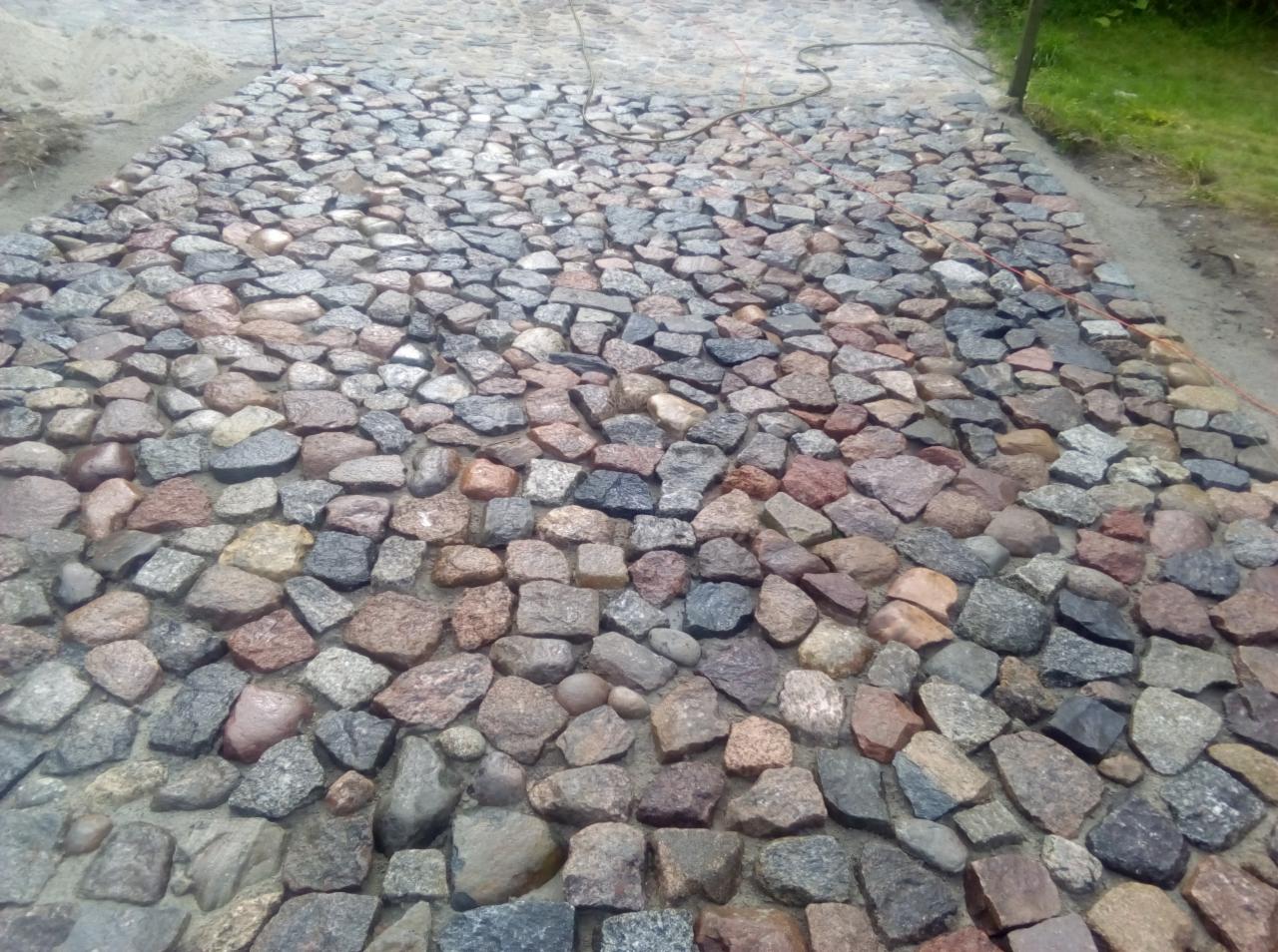 Świeżo ułożona nawierzchnia z kostki granitowej w odcieniach szarości i brązu, widoczne drobne niedociągnięcia w fugach, zarys trawnika w tle.