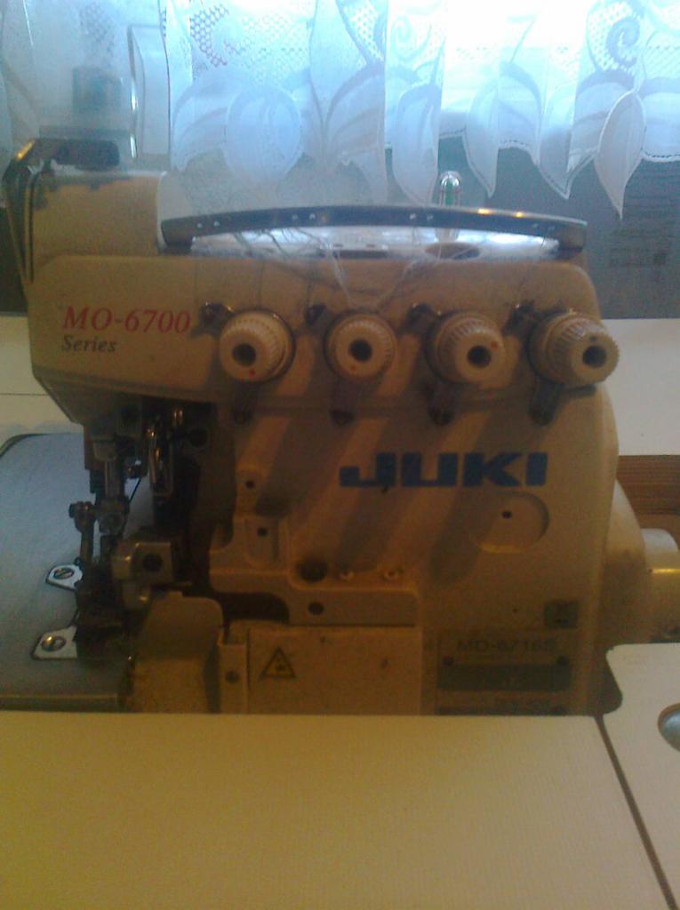 Detale przemysłowej maszyny do szycia JUKI MO-6700 z czterema pokrętłami regulacyjnymi, widoczne fragmenty nici i metalowych elementów, tło stanowi okno z firanką.