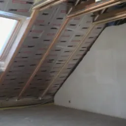 Poddasze w trakcie ocieplania, widoczne skośne ściany z izolacją pokrytą folią, drewniana konstrukcja dachu i okno dachowe, fragment ściany z surowym tynkiem oraz otwór drzwiowy do innego...