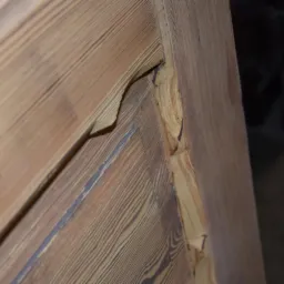 Fragment drewnianej ramy okiennej lub drzwiowej z widocznymi śladami napraw lub renowacji, ukazujący detale uszkodzeń i strukturę drewna.