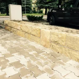 Umyta kostka brukowa przylegająca do niskiego murku z piaskowca, obok zaparkowany samoch&oacute;d.