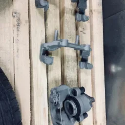 Szare, metalowe zaciski hamulcowe i jarzma po obróbce, leżące na drewnianej palecie, obok opony.
