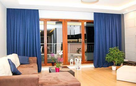 Salon z brązową sofą i dużymi, granatowymi zasłonami na karniszu nad przesuwnymi drzwiami balkonowymi z drewnianą ramą, widok na balkon i kuchnię w tle.