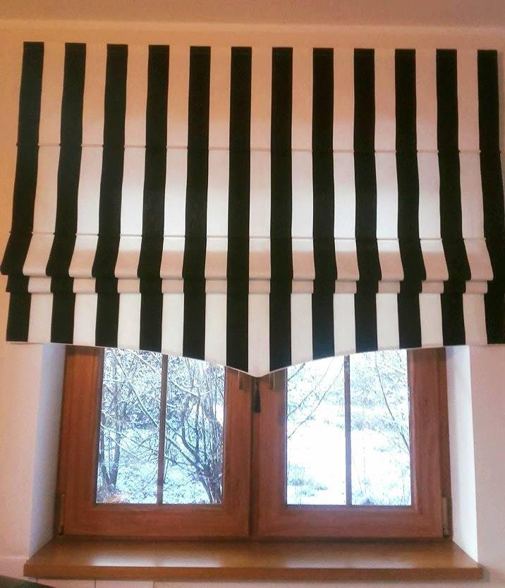 Rzymska roleta w czarno-białe pasy, zamontowana w drewnianym oknie z widokiem na zimowy krajobraz.