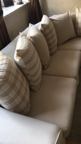 Jasna sofa z beżowymi poduszkami w kratę i paski, ustawionymi pod kątem przy oknie z zasłoną.