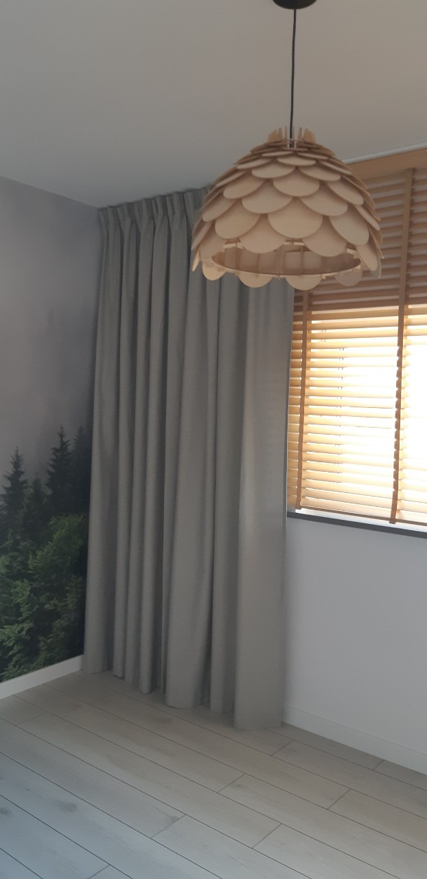 Długie, szare zasłony z marszczeniem przy karniszu, wiszące obok okna z drewnianymi żaluzjami, oświetlone wiszącą lampą z drewnianych elementów, na ścianie fototapeta z motywem lasu.