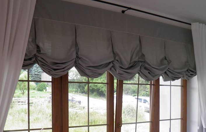 Okno z drewnianą ramą i szarą, drapowaną zasłoną rzymską oraz białymi, półprzezroczystymi firankami po bokach, widok na zieleń za oknem.