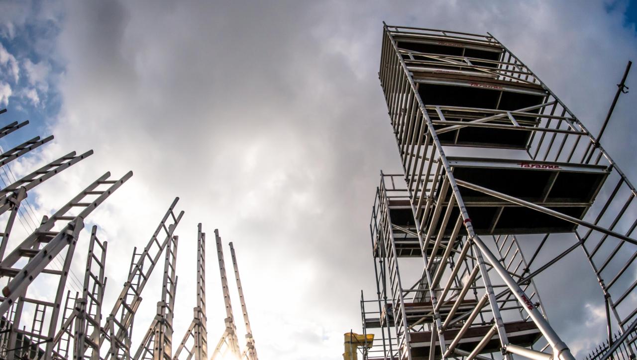 Wysokie, metalowe rusztowania modułowe i drabiny aluminiowe ustawione na placu budowy, widok z dołu na tle pochmurnego nieba.