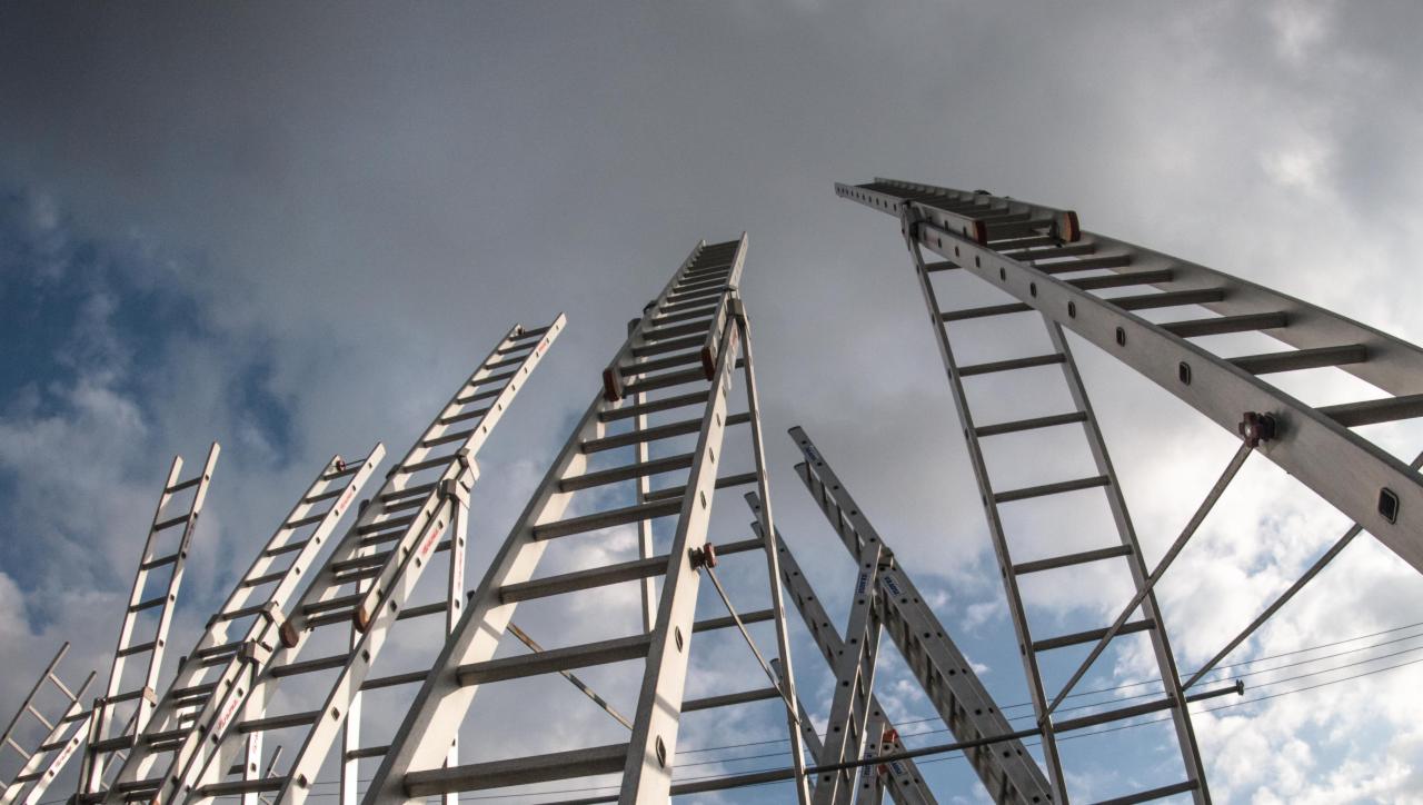 Wiele aluminiowych drabin różnych rozmiarów ustawionych pionowo pod pochmurnym niebem, widok z dołu, widoczne przewody elektryczne w tle.