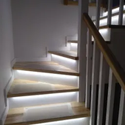 Jasne, drewniane schody z białymi podstopnicami i podświetleniem LED na każdym stopniu; zabezpieczone folią malarską.