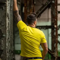 Mężczyzna w żółtej bieliźnie i koszulce, oparty o zniszczoną metalową konstrukcję w opuszczonym budynku.