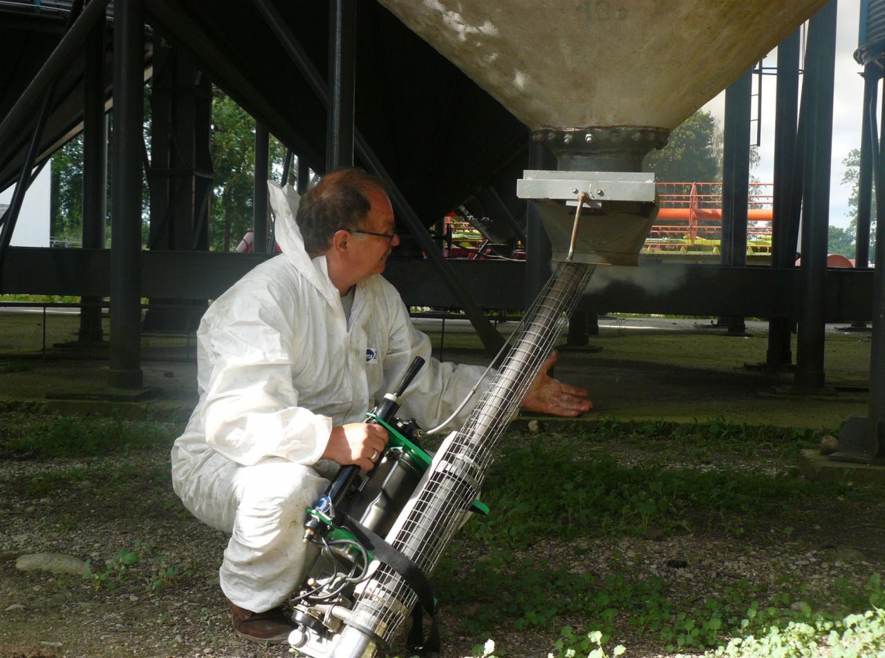 Pracownik w kombinezonie ochronnym, używając zamgławiacza, aplikuje preparat pod silos przemysłowy na zewnątrz.