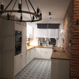 Biała kuchnia w stylu rustykalnym z drewnianym blatem, wzorzystą podłogą i ceglaną ścianą, oświetlona lampą z elementami sznurka.