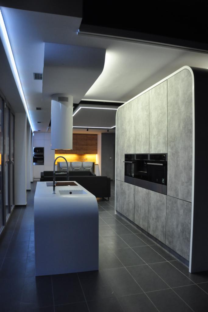 Nowoczesna kuchnia z wyspą i zabudową w odcieniach szarości, minimalistyczny design, oświetlenie LED podkreślające kształty, widok w głąb salonu.