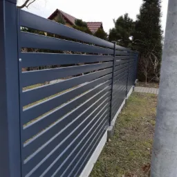 Nowoczesne, poziome ogrodzenie panelowe w kolorze grafitowym, z widoczną podmurówką betonową, na tle domu z czerwonym dachem i zielonych drzew