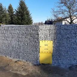 Gabionowe ogrodzenie z żółtą skrzynką gazową na tle drzew i domu, widoczne elementy konstrukcyjne z drutu.