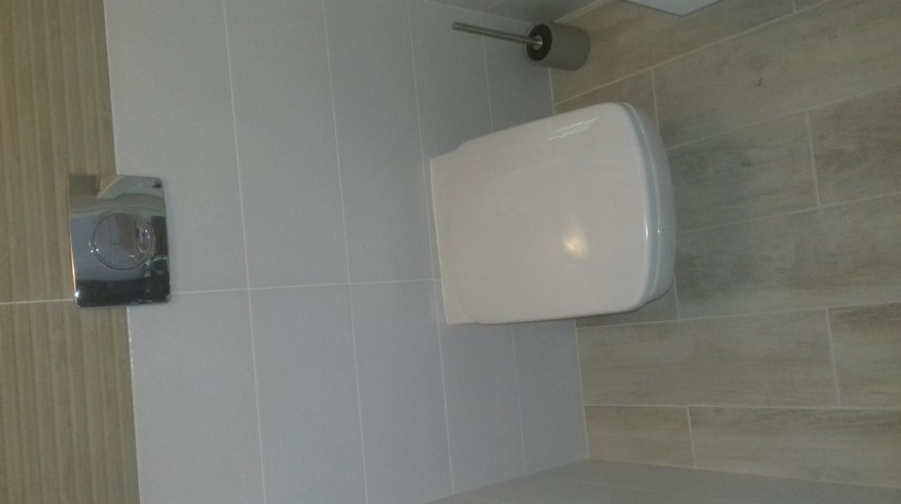 Nowoczesna toaleta wisząca z chromowanym przyciskiem spłukującym i uchwytem na papier toaletowy, wykończona płytkami w odcieniach szarości.