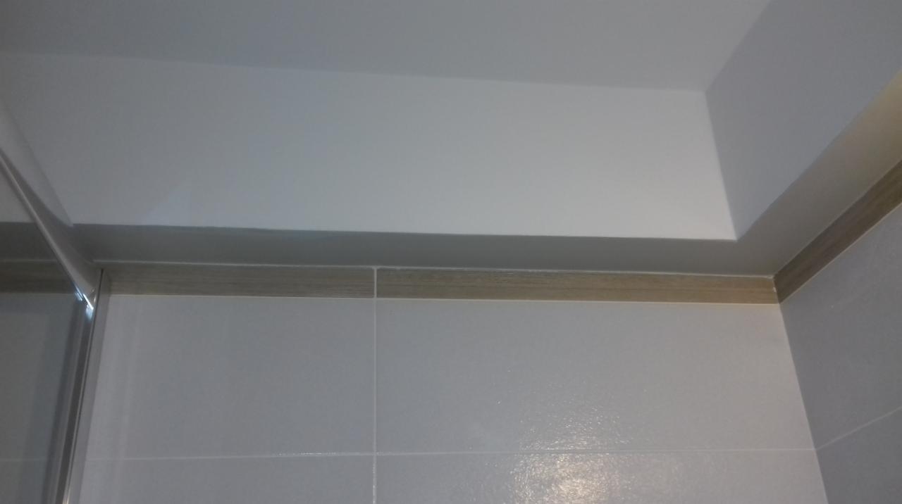 Ściana wyłożona białymi, błyszczącymi płytkami z dekoracyjnym, drewnopodobnym paskiem na styku ze ścianą i sufitem podwieszanym w łazience.