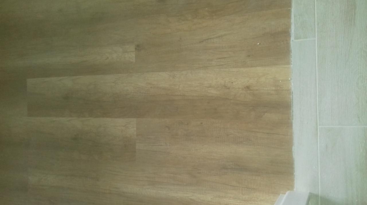 Ułożone panele podłogowe imitujące drewno w odcieniach beżu, sąsiadujące z płytkami ceramicznymi w jasnym odcieniu, widoczne drobne zabrudzenia przy łączeniu.