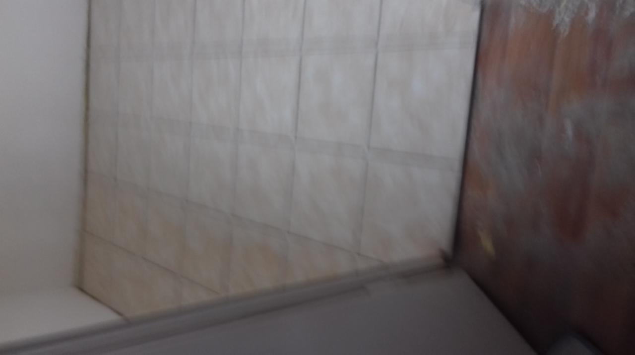 Ściana wyłożona beżowymi płytkami w łazience, widoczne niedokładności w fugowaniu i zabrudzenia. Obok ściana z ciemnego drewna.